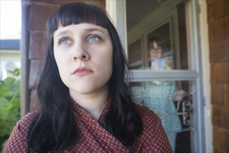 Head and Shoulders Portrait of Contemplative Young Adult Woman with Mid-Adult Woman in Background Looking at her through Front Door