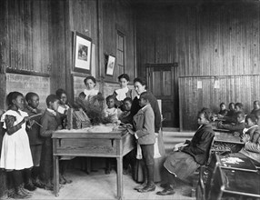 African American Children learning about Thanksgiving, with model Log Cabin on Table, 1900