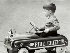 Young boy sitting in toy car with "Fire Chief" written on side