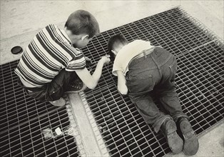 Two Young Boys peering through Subway Grate on Sidewalk while lowering a String with a fastener to snag a Prize