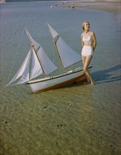 Portrait of Woman in Two-Piece Swimsuit standing on shore near Miniature Sailboat