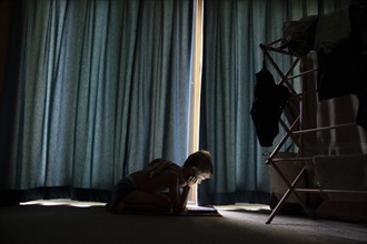 Young boy on floor reading by sunlight seeping through closed curtains