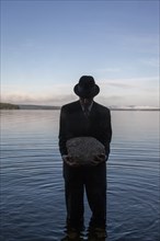 Man in suit and hat holding boulder while standing in calm lake at sunset