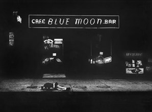 Man lying on sidewalk in front of bar with sign above reading "Cafe Blue Moon Bar"