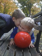 Two young boys being playful with red soccer ball on park bench