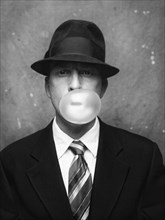 Half-length portrait of mid-adult man in suit and hat blowing a gum bubble