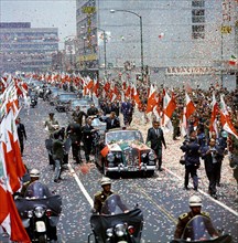 U.S. President John F. Kennedy waving to crowd from convertible car as confetti falls during