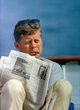 U.S. President John F. Kennedy smoking cigar and reading newspaper while sitting aboard