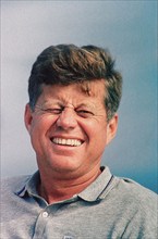 Head and shoulders portrait of squinting and smiling U.S. President John F. Kennedy sitting aboard