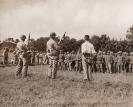 Japanese soldiers line up for formal surrender ceremonies as U.S. Marines with tommy guns and fixed