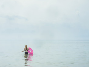 Rear view of young adult woman wading in water holding a pink inner tube