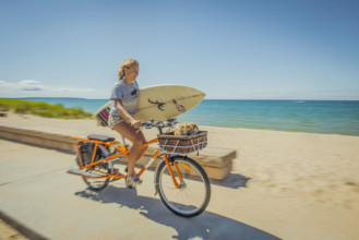 Young teen girl holding surfboard while riding bicycle along beach