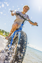 Young teen girl riding bicycle through water at beach