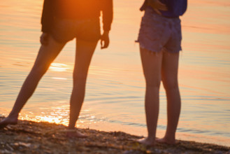 Waist-down portrait of two teen girls standing on shore at sunset