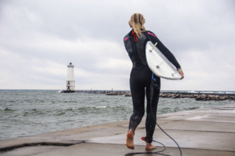 Rear view of young adult woman in wetsuit standing with surf board on Frankfort North lighthouse