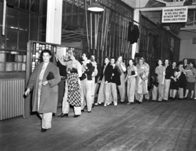 Women war workers wearing slacks punching time cards at end of shift, De Soto bomber plant,