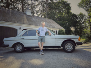 Young adult man with bleached hair leaning against vintage Mercedes car in driveway