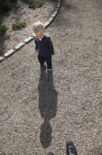 High angle view of young boy casting long shadow on gravel driveway