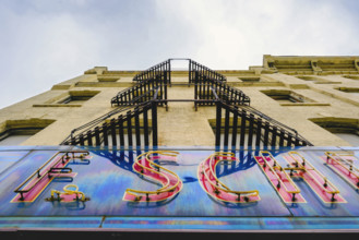 Old neon sign attached to painted brick building with metal balconies, low angle view