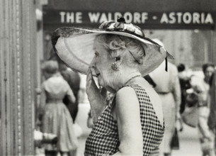 Half-length portrait of elderly woman in wide-brimmed hat standing outside Waldorf-Astoria Hotel,