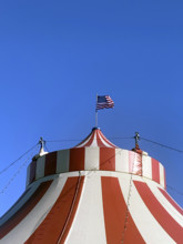Top of red and white circus tent with American flag against blue sky
