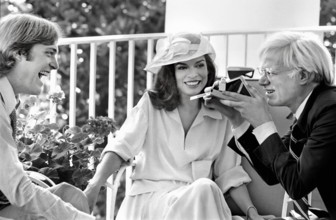 American artist Andy Warhol taking Polaroid picture while sitting with Jack Ford and Bianca Jagger