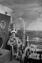 A man in a sailor's cap and eyepatch sits in a reconstructed ship's cabin, c1945-c1965