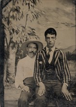 Young Man in a Striped Jacket, Sitting on the Lap of Another Man in Front of Painted Outdoor Backdrop, 1860s-80s.