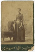 Photograph of a woman standing next to a chair and holding a book, late 19th century. Creator: Louis Janousek.