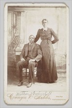 Photograph of a man sitting down with a woman standing next to him, 1880s - 1900. Creator: Cronyn & Hibbs.