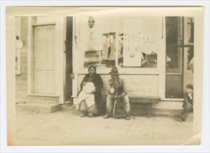 Photograph of a man and woman sitting outside of a storefront, early 20th century. Creator: Unknown.