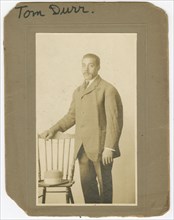 Photograph of Tom Durr standing next to a chair with a hat on it, ca. 1910. Creator: Unknown.