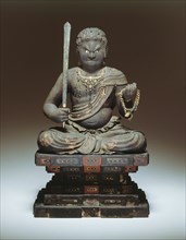 Fudo Myo-o, 13th century. A wooden sculpture of an angry looking diety, holding a sword in his right hand and ope in his left, sitting cross-legged on an stylized rock formation