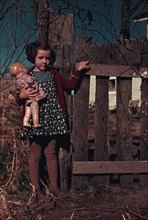 Girl with doll standing by fence, between 1941 and 1942. Creator: Unknown.