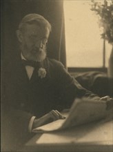 Bearded man in spectacles reading a newspaper, c1900.