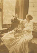 Young woman seated by a window with flowers in her lap, c1900.