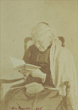 Elderly woman seated and reading a book with knitting in her lap, 1895.