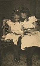 Two girls wearing white dresses and dark stockings reading a book, c1900.