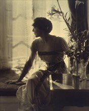Woman sitting on window seat, looking left, full-length portrait, between 1900 and 1920.