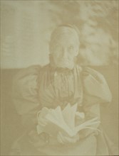 Elderly woman sitting in a chair holding a book, half-length portrait, facing front, c1890.