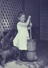 Little girl wearing white chemise, beaded necklace, and high-button shoes on porch with butter churn, c1900.