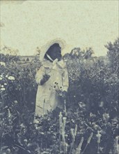 Young African-American(?) girl, wearing long dress and bonnet, standing in field holding flowers, c1900.