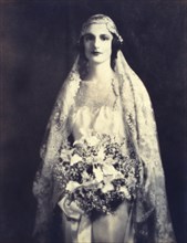 Bride holding a wedding bouquet, facing front, three-quarter length portrait, between 1920 and 1930.