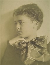 Head-and-shoulders portrait of a young boy with a large plaid bow at the neck of his jacket, facing left, c1900.
