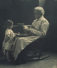 Elderly woman in a rocking chair peeling apple with young girl standing in front of her, c1900.