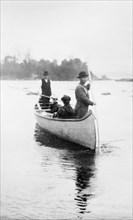 Two Indian guides(?) standing, and two women seated, in canoe, in the Sault Sainte Marie region of Michigan, 1903.