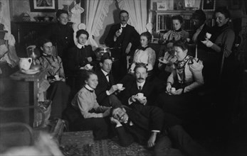 Group portrait of friends at social gathering, seated and standing with tea cups, in domestic setting, between 1890 and 1910.