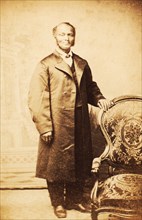 Studio portrait of unidentified man, standing, wearing long coat, c1865. Creator: Sutterley Brothers.