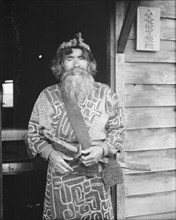Ainu chief wearing a headdress and holding a sword standing in a doorway, 1908. Creator: Arnold Genthe.