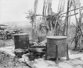 Ainu man seated outdoors on a mat covered with clay containers, 1908. Creator: Arnold Genthe.